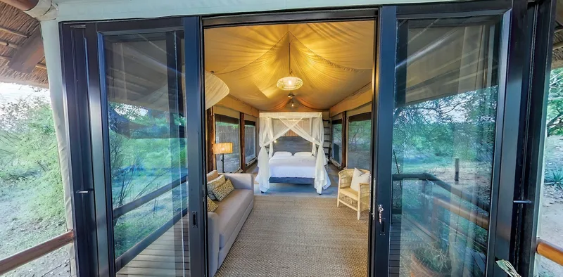 Looking through the open glass doors of a tented suite to the bed inside, lit warmly at dusk