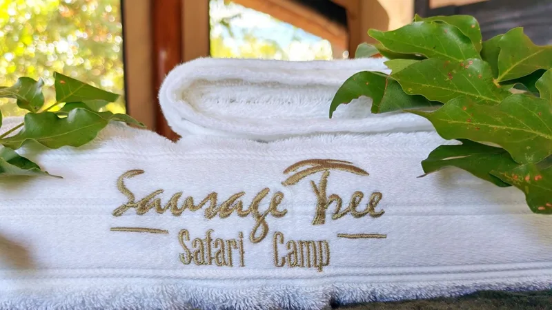 A rolled white towel embroidered with the Sausage Tree Safari Camp name, beside green foliage