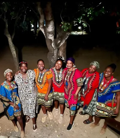 A group of women in brightly coloured traditional dress dancing under the trees